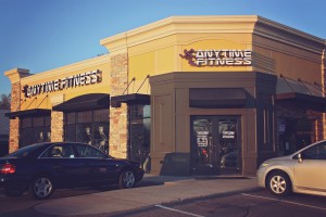 Outside a yellow, grey and brick Anytime Fitness gym with two cars outside