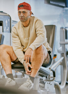 Male with Ta Moko sitting at a leg curl machine inside of anytime fitness wearing a red hat and tan jumper