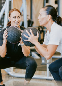 Two women squatting with medicine balls inside of Anytime Fitness